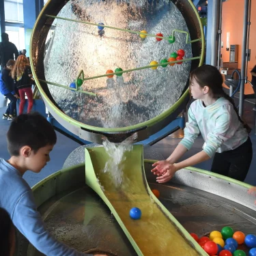 Two kids are playing at the science museum. They are facing a water system machine that contains plastic balls sliding down. They are trying to catch the ball in the water.