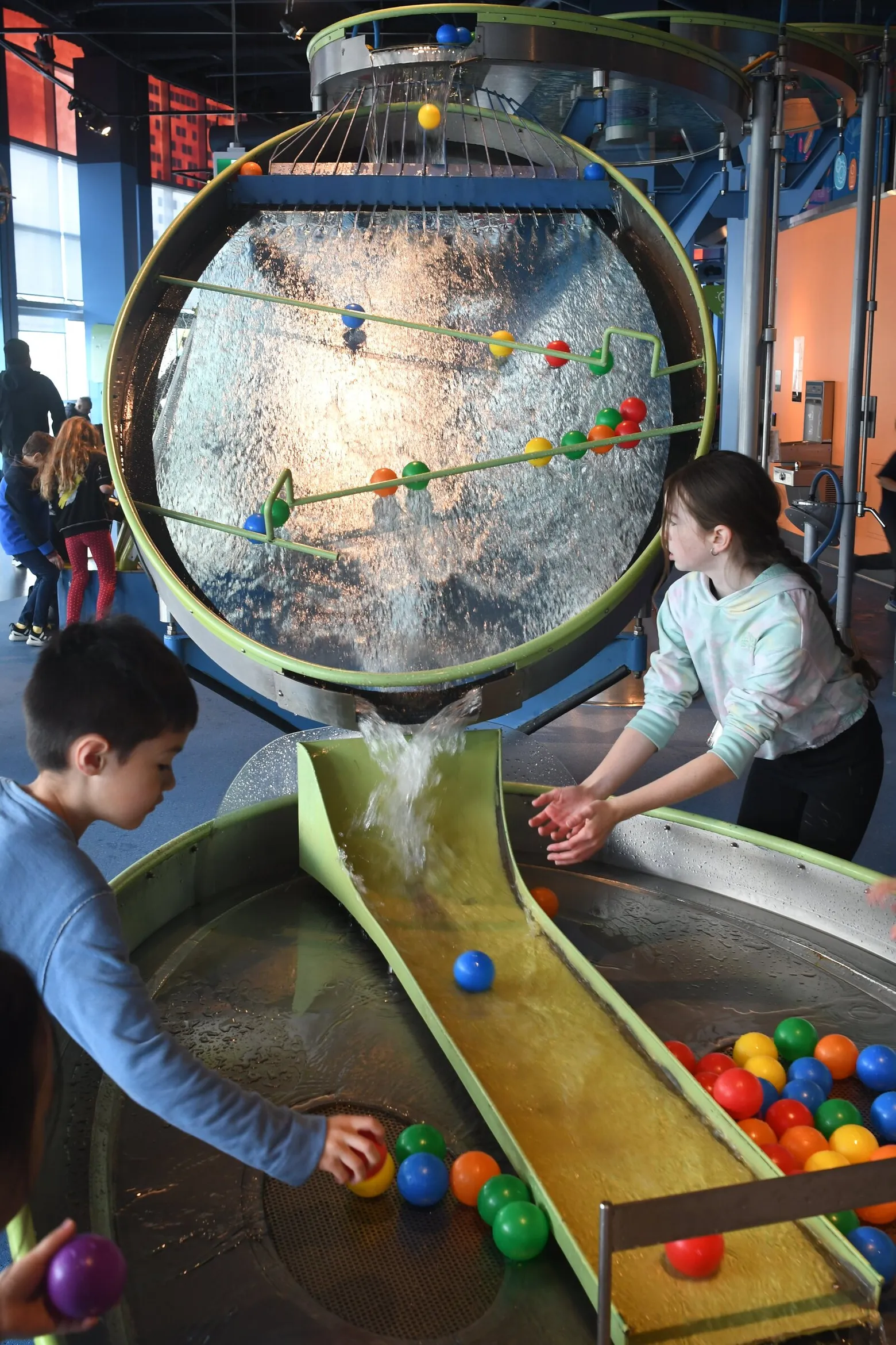 Two kids are playing at the science museum. They are facing a water system machine that contains plastic balls sliding down. They are trying to catch the ball in the water.