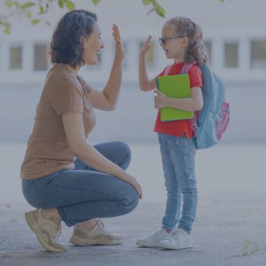 Mother is saying good bye with her loving daughter whcih is around 7 years old. Her daughter is holding a green book and a blue backpack who is going to go to school.