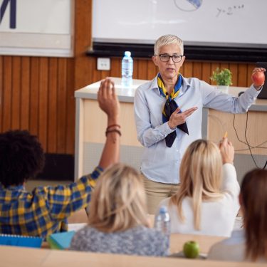 There is a classroom. the teacher is standing in the middle of the classroom, and many students are backward to the camera. the students are hands up to try to answer the question from the teacher, and the teacher is trying to point which student to answer to the question.