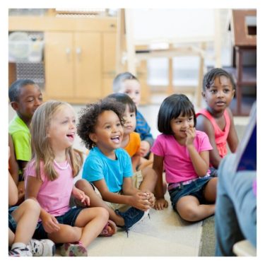 Around 8 children sitting in the middle of the classroom. a teacher is sitting backward to us who is teaching the kids by using a board. the kids are all happily to listen.