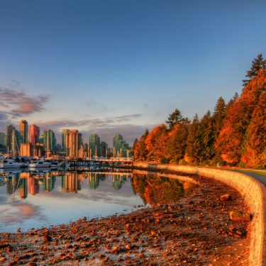 Landscape of a autumn of stanley park. A lake is at the left hand size, a round is at the right hand side, and besides the trees, the sky is blue and orange with a sunset.